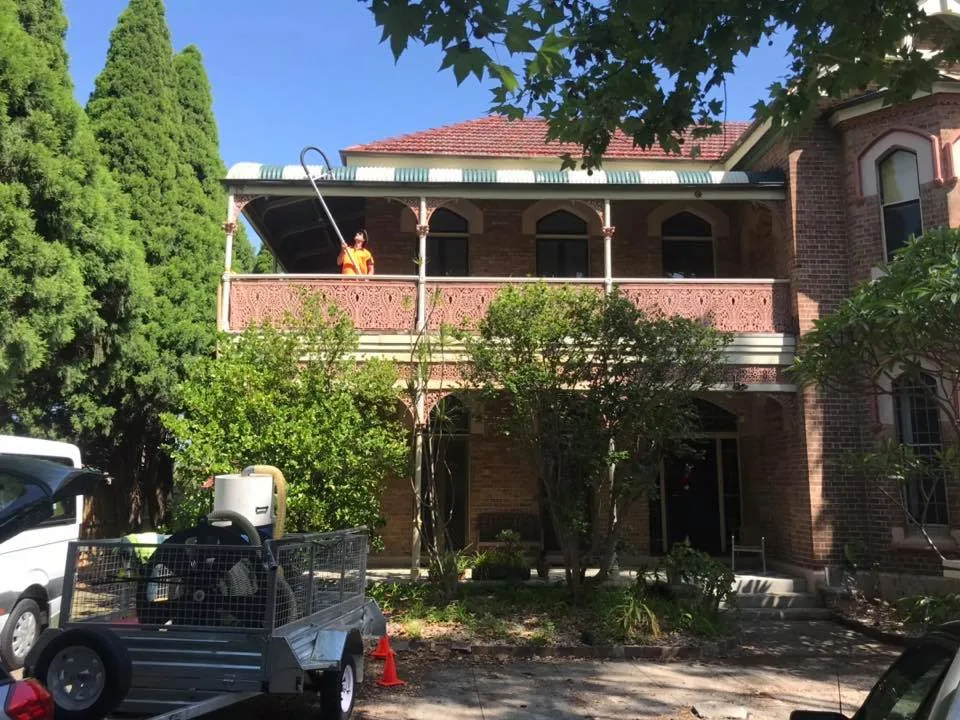 A trailer-mounted gutter vacuum parked at the kerb outside a two-storey brick house, a worker on the balcony reaching the gutter with a long pole